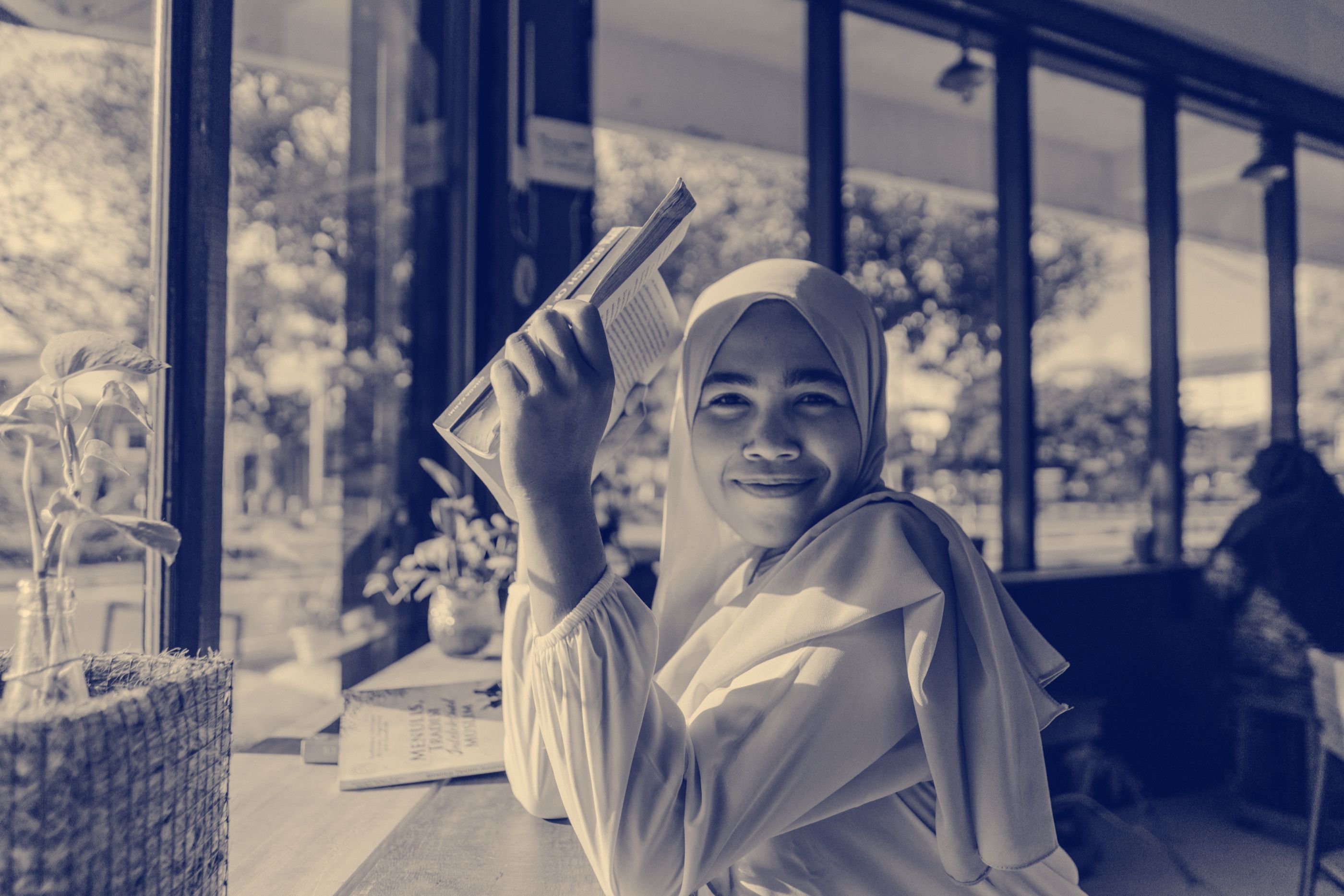 A young woman in a peach hijab smiles in a sunlit window seat, holding a book up beside her head