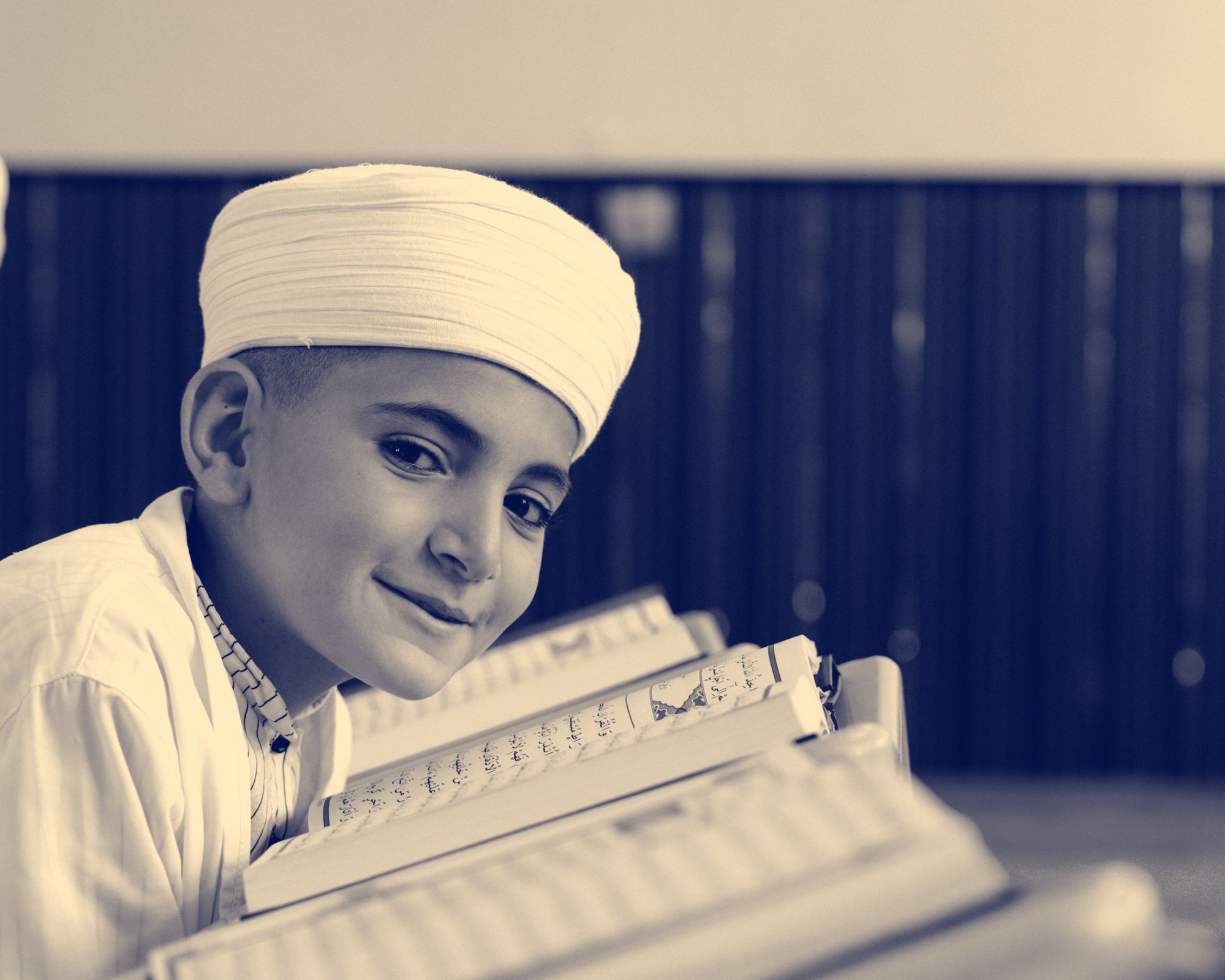 A smiling boy in a white turban and white thobe rests his head beside an open mushaf on a stand