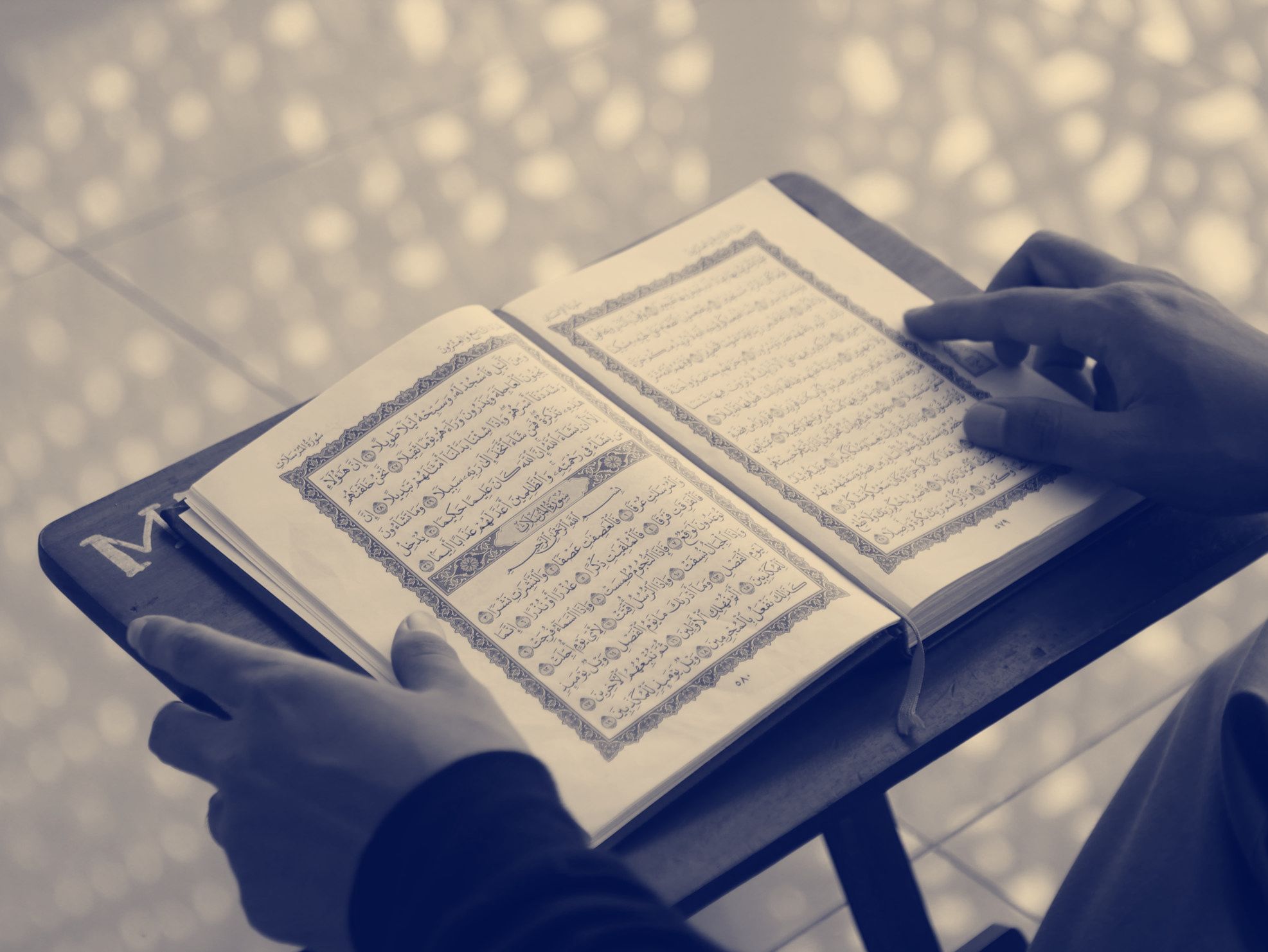 Hands resting on an open mushaf on a wooden stand, one finger following the line