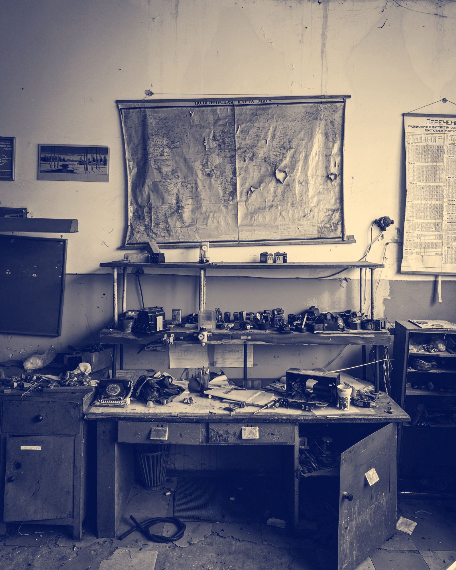 A worn office desk under a faded wall map, with an old rotary phone, loose papers and tools scattered across it